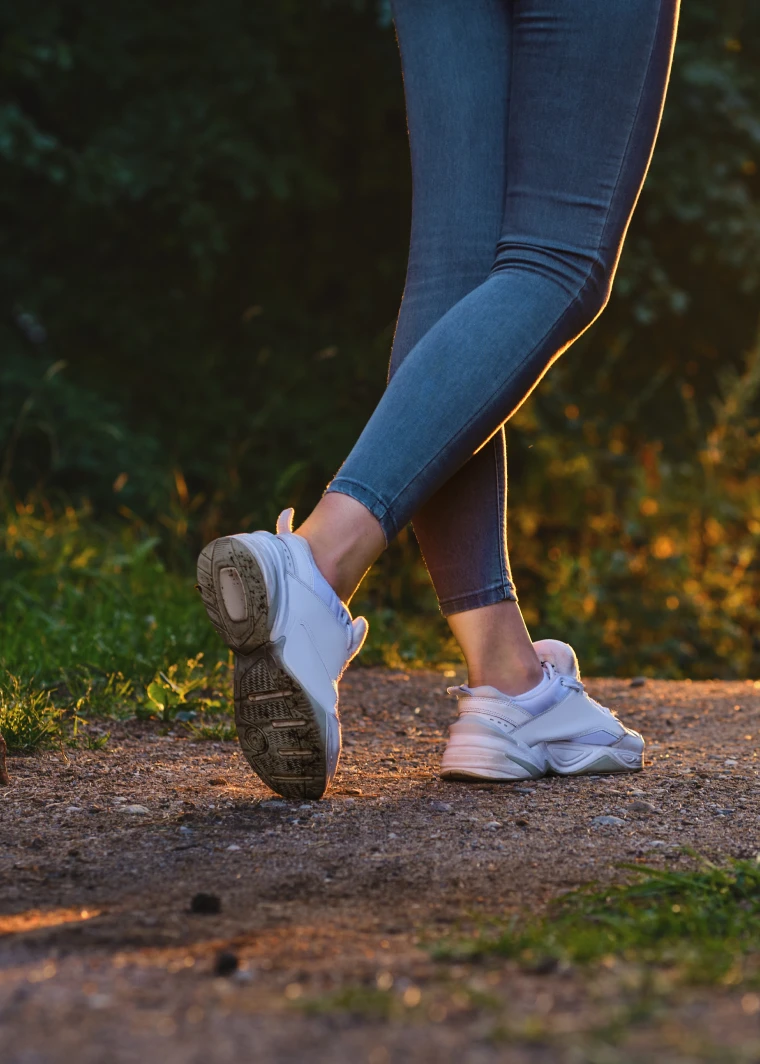 Gambe di una persona con jeans e scarpe sportive bianche mentre cammina su un sentiero sterrato.