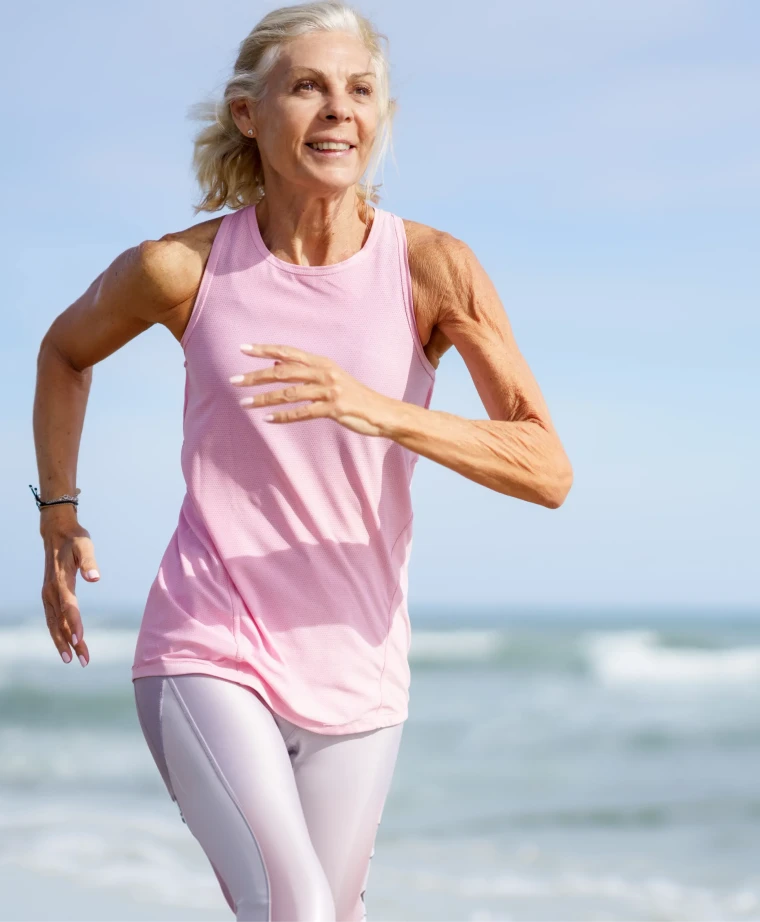 Donna anziana con canotta rosa e leggings chiari mentre corre vicino al mare.