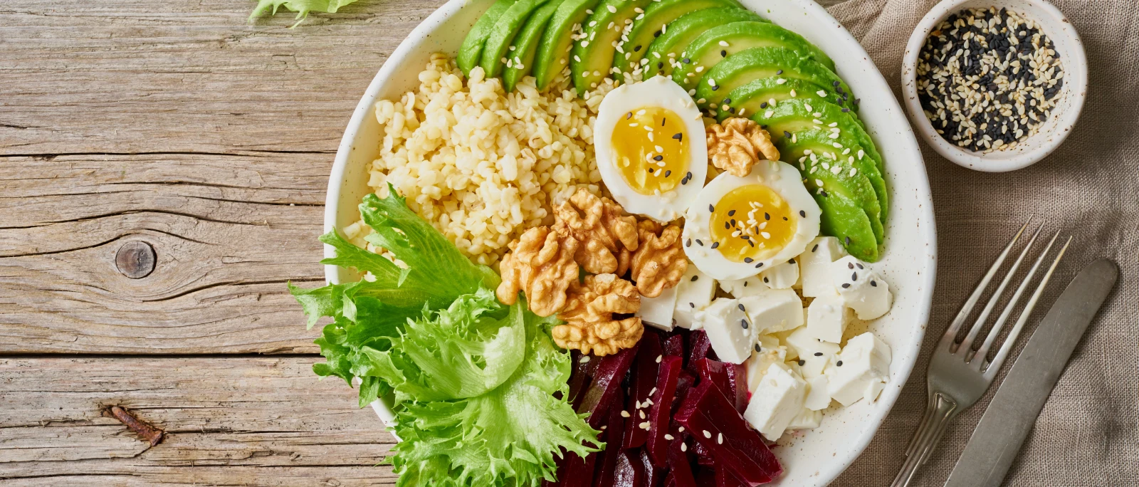 Ciotola con avocado a fette, uova, cereali, formaggio, barbabietole, lattuga e noci su un tavolo di legno.