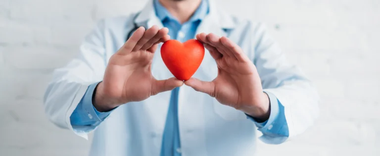 Persona con camice bianco e stetoscopio tiene tra le mani un cuore rosso su sfondo chiaro.