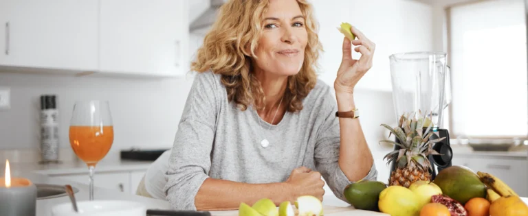 Donna adulta seduta in cucina mentre mangia una fetta di mela, con frutta, frullatore e bicchiere di succo sul piano di lavoro.