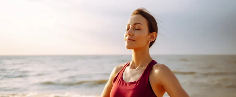 Donna in abbigliamento sportivo in piedi sulla spiaggia, con gli occhi chiusi e le mani sui fianchi, davanti al mare.